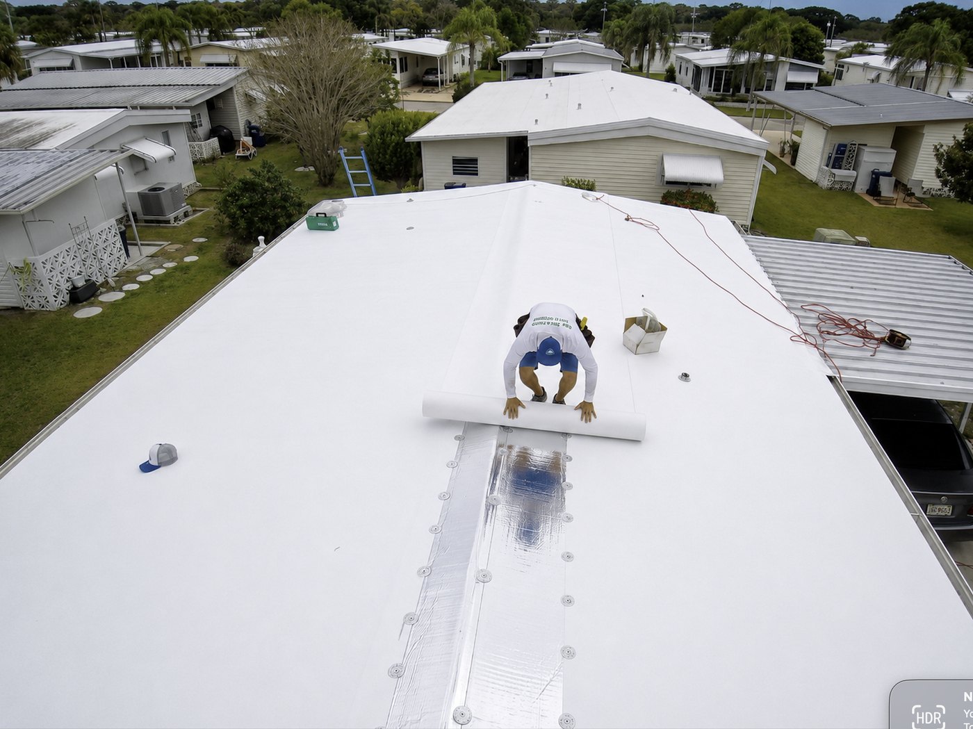 Aerial view of a By The Book Roofing installer rolling out white TPO membrane across the roof of a Florida manufactured home, with the park's other homes visible around it.